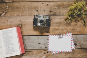 A vintage camera sits on a rustic wooden table next to an open book with a red cover. A bundle of dried yellow flowers is placed to the side, adding to the vintage aesthetic. Two envelopes with airmail design are clipped together next to the camera.
