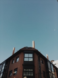 A residential building with a brown brick exterior and multiple windows is viewed from below. Above the building, a clear blue sky stretches across the image, with the moon faintly visible near the upper edge.