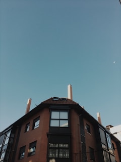 A residential building with a brown brick exterior and multiple windows is viewed from below. Above the building, a clear blue sky stretches across the image, with the moon faintly visible near the upper edge.