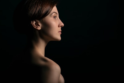 A woman is captured in profile against a dark background. The lighting creates a dramatic effect, highlighting her facial features and casting soft shadows. Her hair is short and styled neatly.