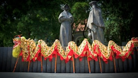 A colorful dragon puppet with a vibrant design is positioned in front of two large stone sculptures of historical figures dressed in traditional robes. The background features lush green trees, and the dragon has patterns of yellow, red, and green, combined with a bright yellow mane and a red-tipped snout.
