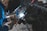 A person wearing protective gloves and a welding helmet is performing welding work, with sparks flying from the equipment. The scene is dimly lit, highlighting the bright blue arc of the welding process.