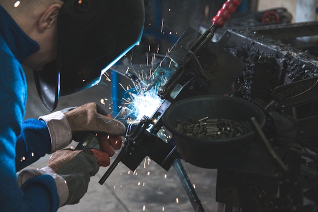 A person wearing protective gloves and a welding helmet is performing welding work, with sparks flying from the equipment. The scene is dimly lit, highlighting the bright blue arc of the welding process.