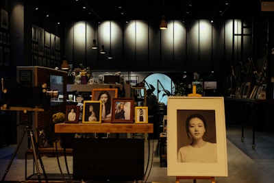 A dimly lit art studio or gallery filled with framed photographs on wooden easels and shelves. A large portrait in the foreground focuses on a serene, expressionless face. Other smaller portraits, mostly of women, are displayed around it. Vintage cameras and decorative plants add to the vintage and artistic ambiance.