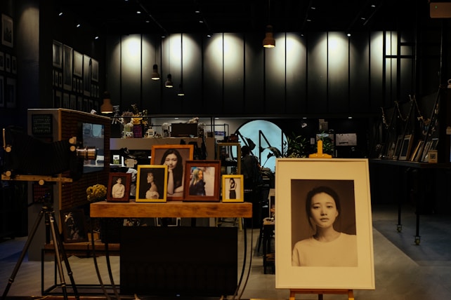 A dimly lit art studio or gallery filled with framed photographs on wooden easels and shelves. A large portrait in the foreground focuses on a serene, expressionless face. Other smaller portraits, mostly of women, are displayed around it. Vintage cameras and decorative plants add to the vintage and artistic ambiance.