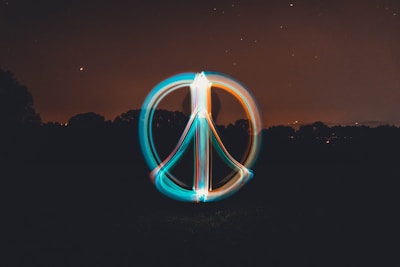 A glowing peace symbol is created using light painting against a dark night sky. The background shows a silhouette of trees with scattered stars and a crescent moon visible in the sky. The symbol is composed of vibrant trails of light in blue and orange colors.