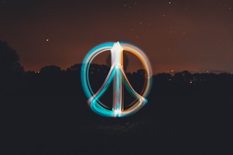 A glowing peace symbol is created using light painting against a dark night sky. The background shows a silhouette of trees with scattered stars and a crescent moon visible in the sky. The symbol is composed of vibrant trails of light in blue and orange colors.