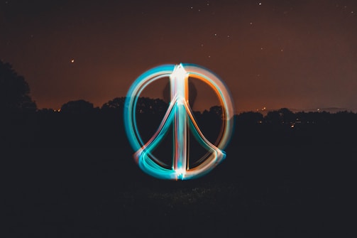 A glowing peace symbol is created using light painting against a dark night sky. The background shows a silhouette of trees with scattered stars and a crescent moon visible in the sky. The symbol is composed of vibrant trails of light in blue and orange colors.