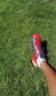 A person wearing a red and black soccer cleat on a lush green grass field. The foot is positioned to the right side of the image, and the grass appears well-maintained.