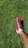 A person wearing a red and black soccer cleat on a lush green grass field. The foot is positioned to the right side of the image, and the grass appears well-maintained.