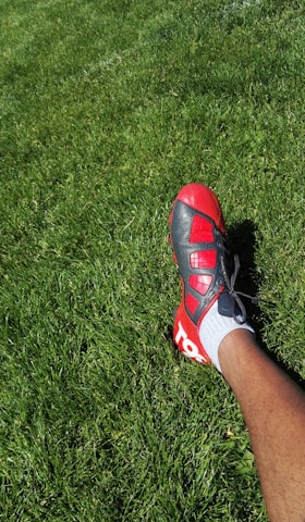 A person wearing a red and black soccer cleat on a lush green grass field. The foot is positioned to the right side of the image, and the grass appears well-maintained.