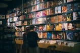 A record store with a wall lined with colorful vinyl albums, featuring various cover art designs and portraits. Two people stand in front of a wooden crate filled with more records, browsing the collection.