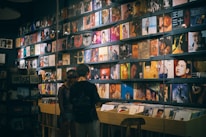 A record store with a wall lined with colorful vinyl albums, featuring various cover art designs and portraits. Two people stand in front of a wooden crate filled with more records, browsing the collection.