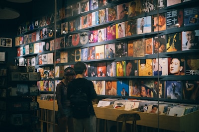 A record store with a wall lined with colorful vinyl albums, featuring various cover art designs and portraits. Two people stand in front of a wooden crate filled with more records, browsing the collection.
