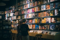 A record store with a wall lined with colorful vinyl albums, featuring various cover art designs and portraits. Two people stand in front of a wooden crate filled with more records, browsing the collection.