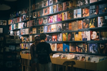 A record store with a wall lined with colorful vinyl albums, featuring various cover art designs and portraits. Two people stand in front of a wooden crate filled with more records, browsing the collection.