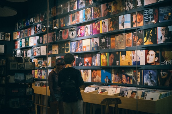A record store with a wall lined with colorful vinyl albums, featuring various cover art designs and portraits. Two people stand in front of a wooden crate filled with more records, browsing the collection.