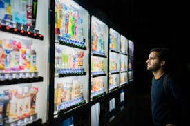 A person standing in front of several illuminated vending machines, examining the selection of drinks and snacks displayed behind the glass panels. The variety includes colorful bottles and packaged food items.