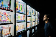 A person standing in front of several illuminated vending machines, examining the selection of drinks and snacks displayed behind the glass panels. The variety includes colorful bottles and packaged food items.