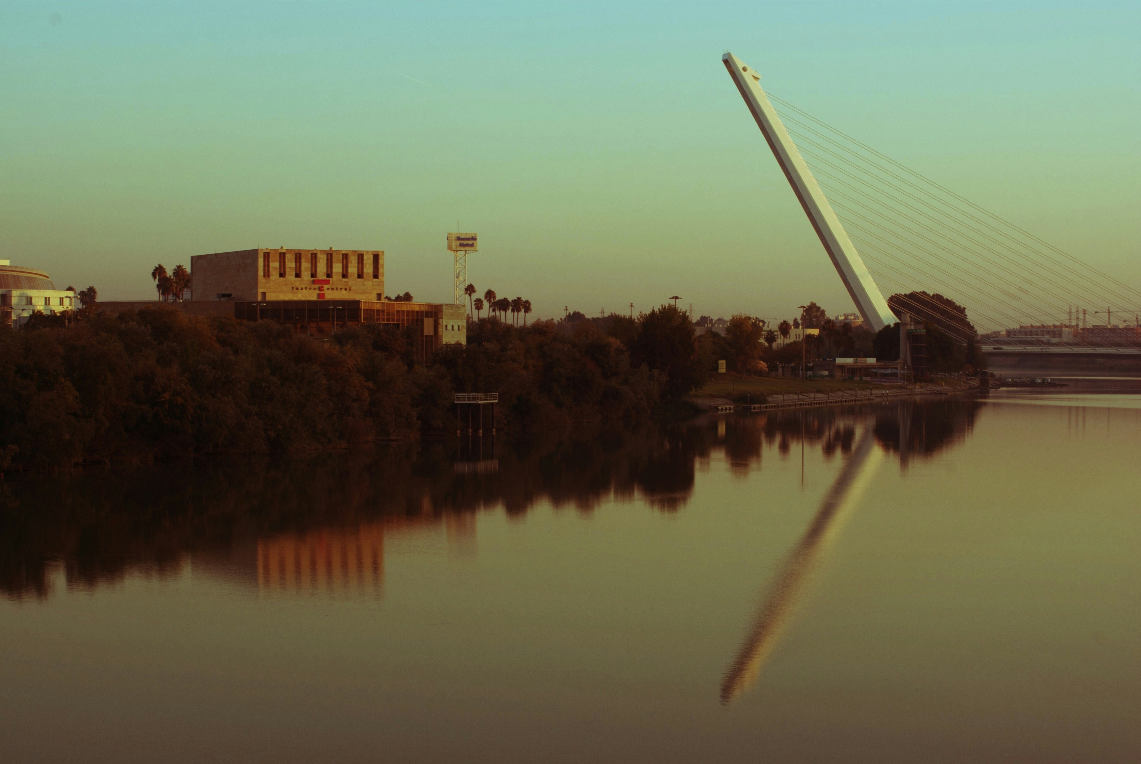 Large suspension bridge over calm water at dusk, steel cables and concrete towers, dim atmospheric lighting, dark water reflections