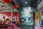 A busy indoor alley or market area with red lanterns hanging from the ceiling. On the right side, a faded yellow and orange cloth hangs near a stack of plastic chairs. On the left, two workers in yellow clothing sit at a table covered with fabric in various shades of pink. In the background, a person is seen crouching on the ground and others are walking further inside the building. Various objects and furniture, including chairs and tables, are scattered throughout the space. The overall setting appears industrial and crowded.