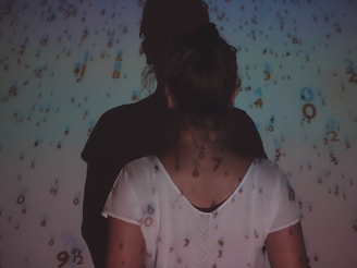 A person wearing a light-colored shirt stands with their back facing the camera, surrounded by projected numbers in various sizes and colors. The image has a mysterious and contemplative atmosphere due to the shadows and abstract numerical overlay.