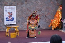 A cultural performance features a performer in an elaborate, colorful costume with intricate designs, standing on a stage. Behind the performer, a poster on the stone wall advertises the Sudbury Dragon Boat Festival. To the right, a vibrant, golden, and orange dragon prop is partially visible, suggesting a traditional dragon dance.