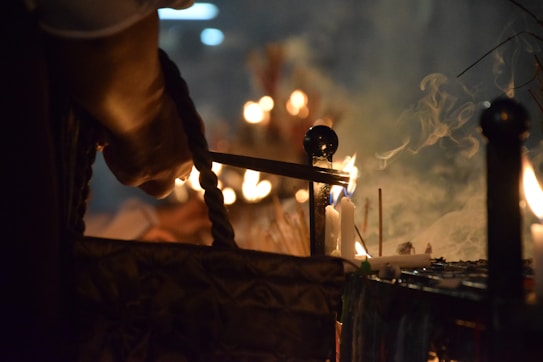 The image captures a close-up of a person lighting candles using a lit taper in a dimly lit setting. Smoke rises up with the soft glow of several lit candles in the background, suggesting a serene or religious atmosphere.