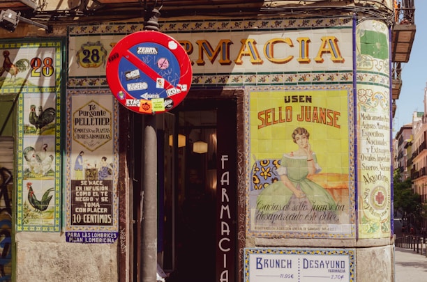 A vintage pharmacy storefront adorned with vibrant ceramic tile artwork featuring advertisements and floral motifs. A 'no entry' street sign covered with stickers is positioned prominently in front of the building.
