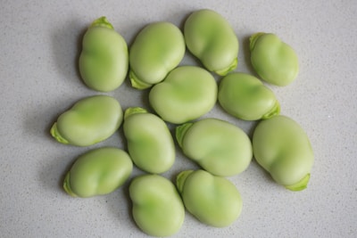 Several light green, smooth beans are scattered on a speckled white surface. The beans are mostly uniform in size and shape, with a few showing parts of a leafy outer shell.