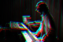 A musician is playing a keyboard or synthesizer in a dimly lit room. The image has a glitch effect with overlapping colors like red, blue, and green, giving a digital, surreal appearance. In the background, there are electronic equipment and a laptop on a table, suggesting a music studio or electronic music setup.