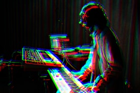 A musician is playing a keyboard or synthesizer in a dimly lit room. The image has a glitch effect with overlapping colors like red, blue, and green, giving a digital, surreal appearance. In the background, there are electronic equipment and a laptop on a table, suggesting a music studio or electronic music setup.