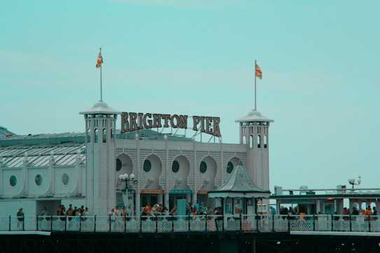 The image features the iconic entrance of Brighton Pier, characterized by its art deco architecture with circular windows and white facade. A large sign reading 'Brighton Pier' is prominently displayed at the top. Crowds of people are visible walking along the pier, and two flags are flying atop the towers.
