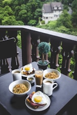 A cozy outdoor setting featuring a table on a balcony with a large breakfast spread consisting of bowls of noodles, plates with eggs on toast, mugs of coffee, and a glass of iced coffee. The background showcases lush green trees and a glimpse of a large house.