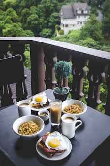A cozy outdoor setting featuring a table on a balcony with a large breakfast spread consisting of bowls of noodles, plates with eggs on toast, mugs of coffee, and a glass of iced coffee. The background showcases lush green trees and a glimpse of a large house.