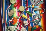 A vibrant collection of Mexican-themed items is arranged on a colorful striped textile. Items include a wooden percussion instrument, a small basket, a red chili pepper, a maraca, a decorative tequila bottle, a clay figurine, a Mexican passport, and several coins. Various ribbons in the colors of the Mexican flag are woven throughout the scene, and a painted gourd with a smiling face adds a whimsical touch.