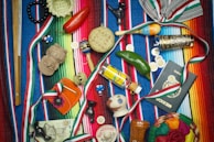 A vibrant collection of Mexican-themed items is arranged on a colorful striped textile. Items include a wooden percussion instrument, a small basket, a red chili pepper, a maraca, a decorative tequila bottle, a clay figurine, a Mexican passport, and several coins. Various ribbons in the colors of the Mexican flag are woven throughout the scene, and a painted gourd with a smiling face adds a whimsical touch.