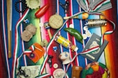 A vibrant collection of Mexican-themed items is arranged on a colorful striped textile. Items include a wooden percussion instrument, a small basket, a red chili pepper, a maraca, a decorative tequila bottle, a clay figurine, a Mexican passport, and several coins. Various ribbons in the colors of the Mexican flag are woven throughout the scene, and a painted gourd with a smiling face adds a whimsical touch.