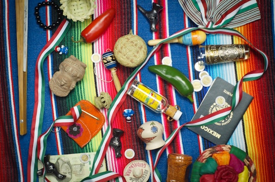A vibrant collection of Mexican-themed items is arranged on a colorful striped textile. Items include a wooden percussion instrument, a small basket, a red chili pepper, a maraca, a decorative tequila bottle, a clay figurine, a Mexican passport, and several coins. Various ribbons in the colors of the Mexican flag are woven throughout the scene, and a painted gourd with a smiling face adds a whimsical touch.