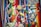 A vibrant collection of Mexican-themed items is arranged on a colorful striped textile. Items include a wooden percussion instrument, a small basket, a red chili pepper, a maraca, a decorative tequila bottle, a clay figurine, a Mexican passport, and several coins. Various ribbons in the colors of the Mexican flag are woven throughout the scene, and a painted gourd with a smiling face adds a whimsical touch.