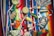 A vibrant collection of Mexican-themed items is arranged on a colorful striped textile. Items include a wooden percussion instrument, a small basket, a red chili pepper, a maraca, a decorative tequila bottle, a clay figurine, a Mexican passport, and several coins. Various ribbons in the colors of the Mexican flag are woven throughout the scene, and a painted gourd with a smiling face adds a whimsical touch.