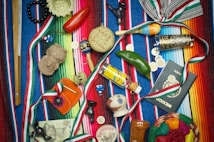 A vibrant collection of Mexican-themed items is arranged on a colorful striped textile. Items include a wooden percussion instrument, a small basket, a red chili pepper, a maraca, a decorative tequila bottle, a clay figurine, a Mexican passport, and several coins. Various ribbons in the colors of the Mexican flag are woven throughout the scene, and a painted gourd with a smiling face adds a whimsical touch.