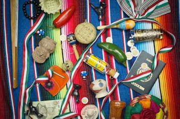 A vibrant collection of Mexican-themed items is arranged on a colorful striped textile. Items include a wooden percussion instrument, a small basket, a red chili pepper, a maraca, a decorative tequila bottle, a clay figurine, a Mexican passport, and several coins. Various ribbons in the colors of the Mexican flag are woven throughout the scene, and a painted gourd with a smiling face adds a whimsical touch.