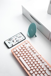 A sleek, modern setup featuring a vintage-style keyboard with rounded keys and a rose gold frame. Nearby, a smartphone displays a gallery of images, and a teal wireless charging stand sits next to them. The background consists of a minimalist white desk.