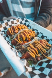 A tray with three tacos is held by a person. The tacos are wrapped in tortillas, filled with a mix of vegetables and meat, and topped with a generous amount of orange sauce. The tray has a black and white checkered paper liner.