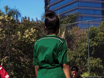 A person wearing a green sports jersey stands on a field. Surrounding the field are lush green trees and a chain-link fence, with a modern glass building visible in the background.