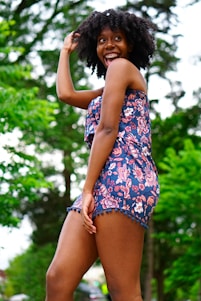 A joyful young woman is outdoors, wearing a floral romper and displaying an expressive, happy demeanor. Her curly hair and vibrant outfit stand out against a backdrop of lush greenery.