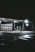 A dimly lit car wash with checkered walls and bright overhead lights. The floor is wet with reflective puddles, and the environment is mostly dark and empty.