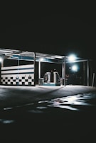 A dimly lit car wash with checkered walls and bright overhead lights. The floor is wet with reflective puddles, and the environment is mostly dark and empty.
