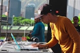 A DJ is focusing on his work at a music event, using a laptop and DJ equipment. He is wearing a mustard-colored long-sleeve shirt and a black cap. In the background, there is a cityscape with tall buildings and green hills.